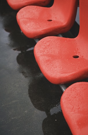 Red plastic chairs with droplets of water on their surface, indicating recent rain. The chairs are arranged in a row and their reflection is visible on the wet ground below.