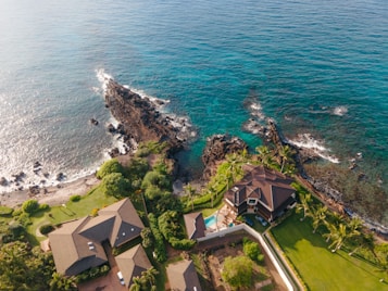 Aerial view of a coastal landscape featuring several upscale houses surrounded by lush greenery. Rock formations extend into the ocean, where waves gently crash against them. The ocean has a rich blue color, complemented by the sandy and rocky shoreline. Palm trees and well-maintained lawns add to the tropical atmosphere.