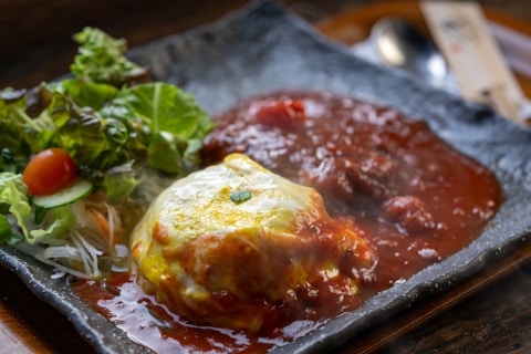 A dish featuring an omelette covered with a rich tomato-based sauce, accompanied by a side salad with lettuce, cherry tomatoes, and cucumber slices. The meal is served on a textured ceramic plate.