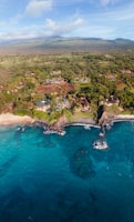 An expansive coastline with clear turquoise waters and rocky formations meeting a lush, green landscape. Scattered houses and villas are nestled among the trees, with mountains visible in the distance under a partly cloudy sky.