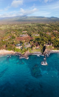 An expansive coastline with clear turquoise waters and rocky formations meeting a lush, green landscape. Scattered houses and villas are nestled among the trees, with mountains visible in the distance under a partly cloudy sky.