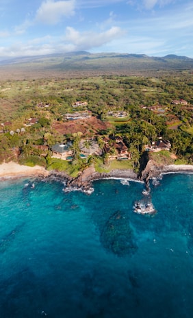An expansive coastline with clear turquoise waters and rocky formations meeting a lush, green landscape. Scattered houses and villas are nestled among the trees, with mountains visible in the distance under a partly cloudy sky.