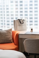 A shopping bag with the words 'Golden Goose' is placed on a light wooden desk next to a grey chair. An orange chair with a grey cushion is positioned in the foreground, and the background shows a blurry urban view through a large window.