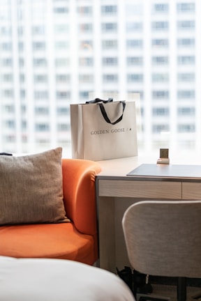 A shopping bag with the words 'Golden Goose' is placed on a light wooden desk next to a grey chair. An orange chair with a grey cushion is positioned in the foreground, and the background shows a blurry urban view through a large window.