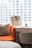 A shopping bag with the words 'Golden Goose' is placed on a light wooden desk next to a grey chair. An orange chair with a grey cushion is positioned in the foreground, and the background shows a blurry urban view through a large window.