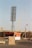 A large metal floodlight tower is positioned in front of a stadium with curved seating structures. A red and white striped barrier is in the foreground, alongside a small red building with a brown base. A tall, yellow metal framework crane extends diagonally across the scene.