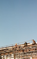 Several construction workers are sitting and working on a high scaffold structure with clear blue sky in the background. They appear to be wearing safety gear, including helmets and harnesses, while surrounded by a network of metal beams.