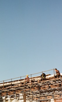 Several construction workers are sitting and working on a high scaffold structure with clear blue sky in the background. They appear to be wearing safety gear, including helmets and harnesses, while surrounded by a network of metal beams.