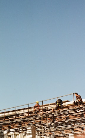 Several construction workers are sitting and working on a high scaffold structure with clear blue sky in the background. They appear to be wearing safety gear, including helmets and harnesses, while surrounded by a network of metal beams.