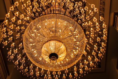 An ornate chandelier with numerous crystal elements and warm glowing lights hangs from the ceiling. The design includes intricate detailing and a symmetrical pattern, creating a luxurious and elegant appearance against a richly decorated ceiling.