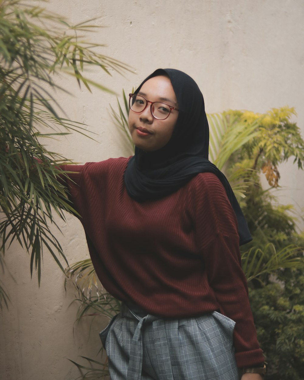 a woman wearing a hijab and glasses standing in front of a plant