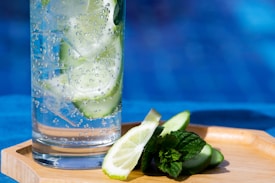 A refreshing glass of sparkling water filled with ice and cucumber slices stands on a wooden tray. Next to the glass, there are slices of cucumber, mint leaves, and a wedge of lime. The background is a vibrant blue, suggesting a sunny day by a pool.