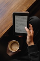 A person is holding an e-reader device in one hand, reading text on the screen, while also holding a mug of coffee in the other hand. The setting appears cozy with a wooden table in the background and the person wearing a dark-colored outfit.