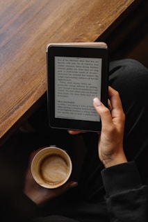 A person is holding an e-reader device in one hand, reading text on the screen, while also holding a mug of coffee in the other hand. The setting appears cozy with a wooden table in the background and the person wearing a dark-colored outfit.