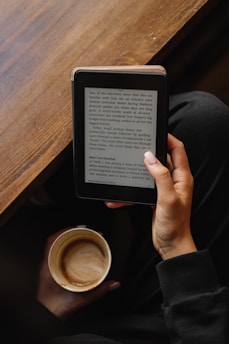 A person is holding an e-reader device in one hand, reading text on the screen, while also holding a mug of coffee in the other hand. The setting appears cozy with a wooden table in the background and the person wearing a dark-colored outfit.