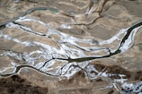 An aerial view of a landscape featuring a network of winding rivers cutting through sandy and rocky terrain. The rivers are partially frozen, with snow or ice accentuating the riverbanks. The land around the rivers appears arid with patches of brown and sparse vegetation.