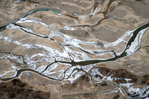 An aerial view of a landscape featuring a network of winding rivers cutting through sandy and rocky terrain. The rivers are partially frozen, with snow or ice accentuating the riverbanks. The land around the rivers appears arid with patches of brown and sparse vegetation.