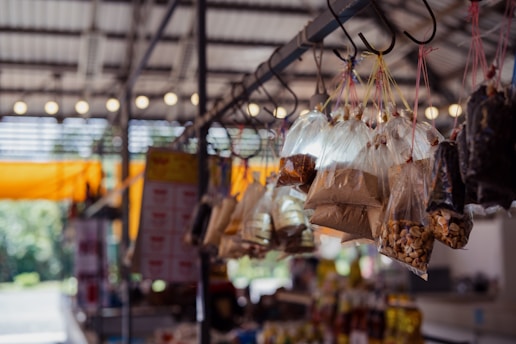 Plastic bags filled with various dry goods hang from hooks in an indoor market setting. The background is slightly blurred, showing other market stalls under a roof with lighting.