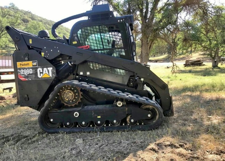 A black CAT 299D compact track loader is stationary on a grassy area. It features a rugged design with visible tracks and construction markings. The background includes green trees and a hilly landscape, suggesting a rural or outdoor setting.
