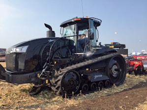 A large black agricultural tractor is parked on a field with a clear blue sky in the background. The vehicle has a massive tracked base and is equipped with modern technology. Nearby, a man is seated in the cabin operating the machine.