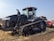 A large black agricultural tractor is parked on a field with a clear blue sky in the background. The vehicle has a massive tracked base and is equipped with modern technology. Nearby, a man is seated in the cabin operating the machine.