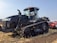 A large black agricultural tractor is parked on a field with a clear blue sky in the background. The vehicle has a massive tracked base and is equipped with modern technology. Nearby, a man is seated in the cabin operating the machine.