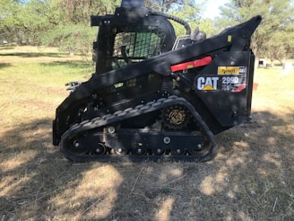 A black compact track loader is parked on grass in an outdoor setting, surrounded by trees and a clear sky. The machine has visible branding and a rugged structure with tracks for movement.
