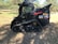 A black compact track loader is parked on grass in an outdoor setting, surrounded by trees and a clear sky. The machine has visible branding and a rugged structure with tracks for movement.