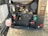 An exposed engine compartment of a vehicle includes various components like hoses, filters, and a battery. The machinery appears well-maintained, with tubes and wires organized, and features various colors like red, green, and black. The ground beneath is dry and grassy.