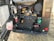 An exposed engine compartment of a vehicle includes various components like hoses, filters, and a battery. The machinery appears well-maintained, with tubes and wires organized, and features various colors like red, green, and black. The ground beneath is dry and grassy.
