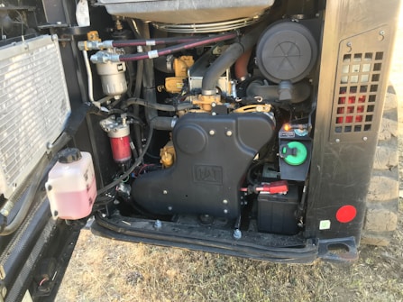 An exposed engine compartment of a vehicle includes various components like hoses, filters, and a battery. The machinery appears well-maintained, with tubes and wires organized, and features various colors like red, green, and black. The ground beneath is dry and grassy.