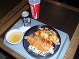 A meal is served on a tray, featuring breaded and fried chicken pieces garnished with cherry tomatoes and green peas. The plate also includes a dish topped with a brown sauce, likely curry or gravy. Accompanying the meal is a cup of soup or broth, a can of Diet Coke, and a larger cup of Coca-Cola, identifiable by the logos and colors.