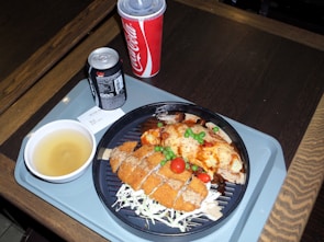 A meal is served on a tray, featuring breaded and fried chicken pieces garnished with cherry tomatoes and green peas. The plate also includes a dish topped with a brown sauce, likely curry or gravy. Accompanying the meal is a cup of soup or broth, a can of Diet Coke, and a larger cup of Coca-Cola, identifiable by the logos and colors.
