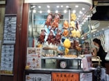 A shop display features various roasted and cooked poultry hanging, including ducks and chickens. The display is encased in glass and illuminated by bright lights. A person behind the counter is serving food to customers, with visible menus and signs featuring Chinese text. The surrounding area shows more menu items and a couple of customers.