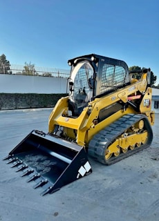 A bright yellow compact track loader with a front bucket attachment is parked on a paved surface. The machine features rubber tracks and a cab with protective metal grating. In the background, there's a white wall adorned with fencing and greenery.