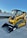A bright yellow compact track loader with a front bucket attachment is parked on a paved surface. The machine features rubber tracks and a cab with protective metal grating. In the background, there's a white wall adorned with fencing and greenery.