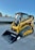 A bright yellow compact track loader with a front bucket attachment is parked on a paved surface. The machine features rubber tracks and a cab with protective metal grating. In the background, there's a white wall adorned with fencing and greenery.