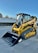A bright yellow compact track loader with a front bucket attachment is parked on a paved surface. The machine features rubber tracks and a cab with protective metal grating. In the background, there's a white wall adorned with fencing and greenery.