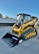 A bright yellow compact track loader with a front bucket attachment is parked on a paved surface. The machine features rubber tracks and a cab with protective metal grating. In the background, there's a white wall adorned with fencing and greenery.