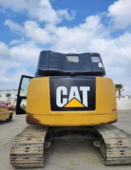 A large construction vehicle, specifically an excavator, is seen from the rear with a prominent 'CAT' logo displayed on the yellow body. The vehicle rests on wide, tracked wheels, and one side door is slightly open. The background includes a blue sky with scattered white clouds, some palm trees, and a structure.