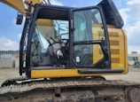 A yellow excavator with a closed cabin and visible control seat, set in an outdoor industrial environment. The machinery sits on a track, indicating it is designed for movement over rough terrain. The surrounding area includes some industrial buildings and equipment.