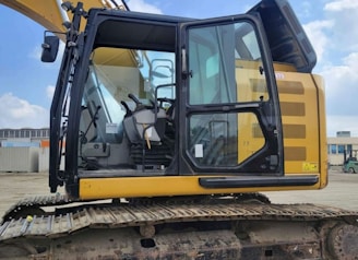 A yellow excavator with a closed cabin and visible control seat, set in an outdoor industrial environment. The machinery sits on a track, indicating it is designed for movement over rough terrain. The surrounding area includes some industrial buildings and equipment.
