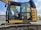 A yellow excavator with a closed cabin and visible control seat, set in an outdoor industrial environment. The machinery sits on a track, indicating it is designed for movement over rough terrain. The surrounding area includes some industrial buildings and equipment.