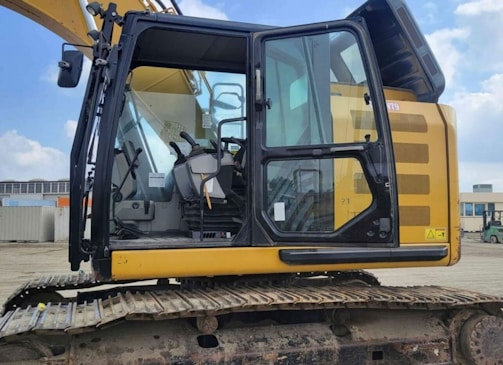 A yellow excavator with a closed cabin and visible control seat, set in an outdoor industrial environment. The machinery sits on a track, indicating it is designed for movement over rough terrain. The surrounding area includes some industrial buildings and equipment.