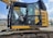 A yellow excavator with a closed cabin and visible control seat, set in an outdoor industrial environment. The machinery sits on a track, indicating it is designed for movement over rough terrain. The surrounding area includes some industrial buildings and equipment.