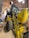 A large yellow backhoe loader with the branding 'New Holland' parked inside a spacious wooden shed. The machine's rear bucket is raised, revealing the detailed mechanical parts. The shed has a curved roof with metal beams and bright fluorescent lights hanging, providing ample illumination. On the left side, there are a few storage items including a bin and a broom.