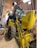 A large yellow backhoe loader with the branding 'New Holland' parked inside a spacious wooden shed. The machine's rear bucket is raised, revealing the detailed mechanical parts. The shed has a curved roof with metal beams and bright fluorescent lights hanging, providing ample illumination. On the left side, there are a few storage items including a bin and a broom.