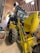 A large yellow backhoe loader with the branding 'New Holland' parked inside a spacious wooden shed. The machine's rear bucket is raised, revealing the detailed mechanical parts. The shed has a curved roof with metal beams and bright fluorescent lights hanging, providing ample illumination. On the left side, there are a few storage items including a bin and a broom.