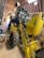 A large yellow backhoe loader with the branding 'New Holland' parked inside a spacious wooden shed. The machine's rear bucket is raised, revealing the detailed mechanical parts. The shed has a curved roof with metal beams and bright fluorescent lights hanging, providing ample illumination. On the left side, there are a few storage items including a bin and a broom.