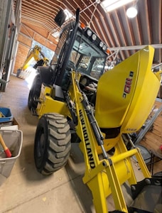 A large yellow backhoe loader with the branding 'New Holland' parked inside a spacious wooden shed. The machine's rear bucket is raised, revealing the detailed mechanical parts. The shed has a curved roof with metal beams and bright fluorescent lights hanging, providing ample illumination. On the left side, there are a few storage items including a bin and a broom.